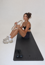 A woman sits on a black yoga mat holding a gymnastic ring, wearing a black sports bra, dark shorts, white socks, and chunky silver sneakers. Two small dumbbells are placed on the mat, which has the brand name 'YÓLA SCULPT' printed on it, against a plain white studio background.