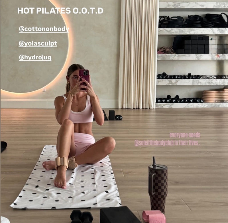 A woman in a pink workout outfit sits cross-legged on a white patterned yoga mat in a fitness studio, holding a phone to take a mirror selfie. The room has wood floors, a circular wall light, shelving with dumbbells, and exercise accessories including pink foam rollers, black blocks, and a patterned tumbler.