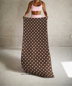 A woman stands in a studio holding up a long, narrow fabric item with a brown background and evenly spaced white polka dots. She is wearing a pink sports bra and matching high-waisted leggings.